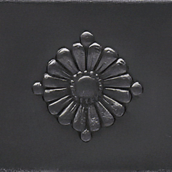 Detailaufnahme eines Ornaments, das den ansonsten schlichten Charakter des Ofens auflockert. Es handelt sich um ein rundes Muster, das eine stilisierte Blüte darstellt.