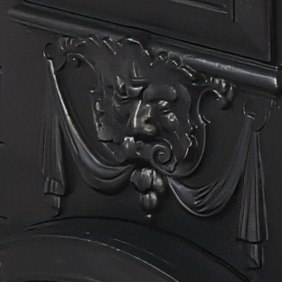 Detailaufnahme des Ornaments über der Brennraumtür: Es zeigt ein mit Blättern und Ranken angedeutetes Gesicht, aus dessen Mund sich zu beiden Seiten Gardinen über die weiterführende Verzierung des Ofens ausbreiten.