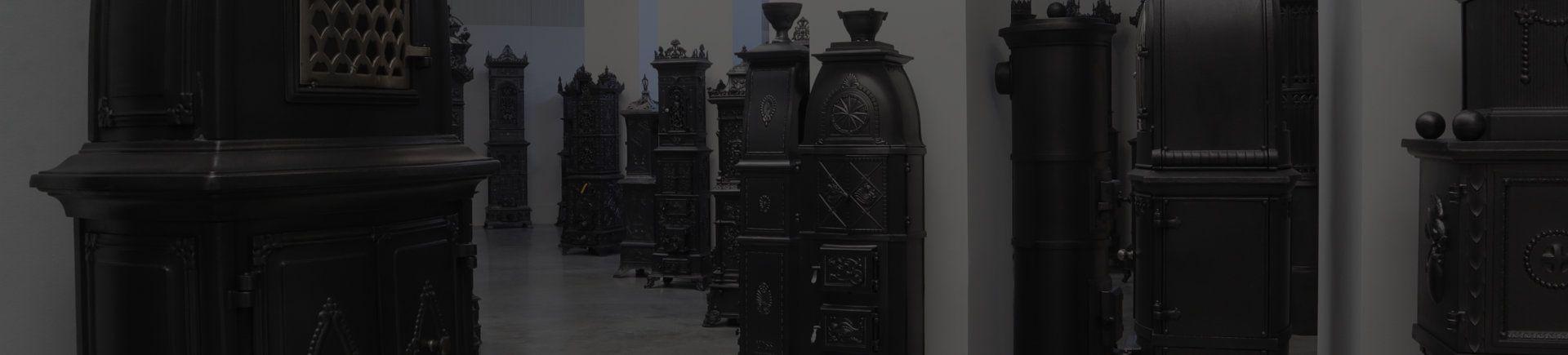 Ausstellung der Antik Ofen Galerie. Zahlreiche antike gusseiserne Öfen sind in Blöcken von vier Stück ausgestellt. Sie reihen sich jeweils um eine Säule auf. 
