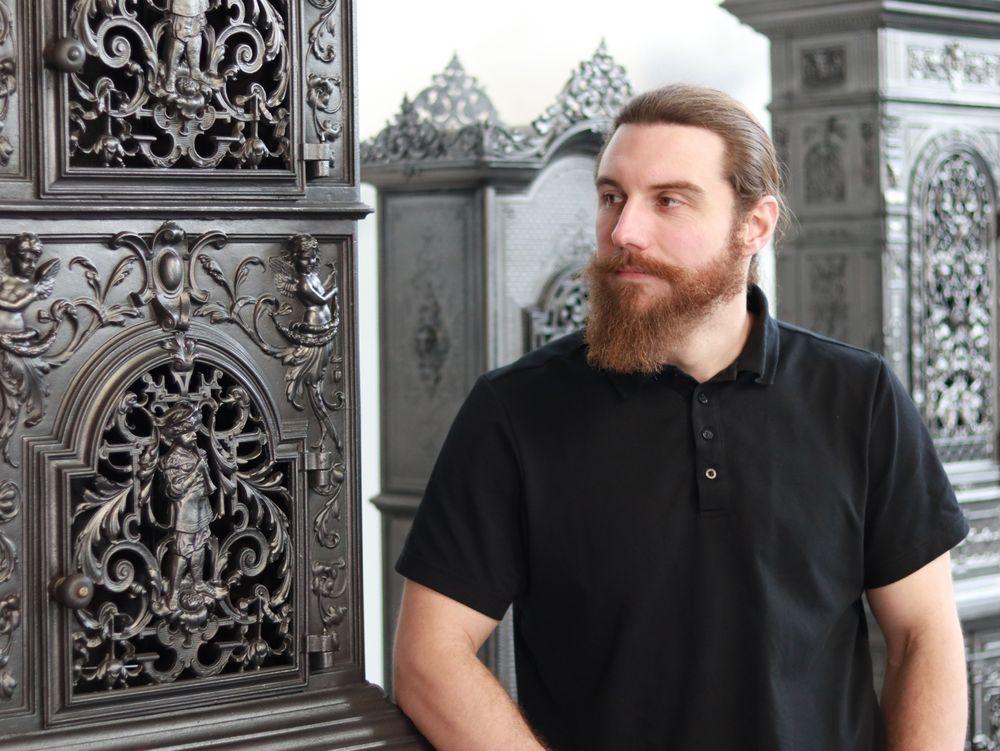 Phillip Neumann, Geschäftsführer der Antik Ofen Galerie, steht vor einem gusseisernen Ofen. Im Hintergrund sind weitere Öfen zu sehen. 